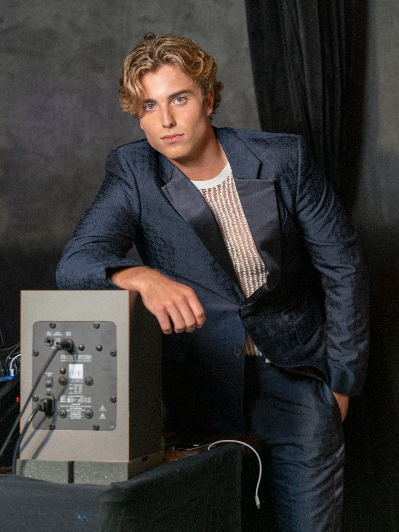 Man in a suit leans on a speaker while making eye contact with the camera